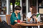 Two women with black hair wearing green and white shirt sitting at table in a street cafe, holding digital tablet, smiling.