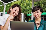 Two women with black hair wearing green and white shirt sitting in front of laptop at table in a street cafe, laughing.