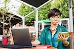 Woman with black hair wearing green shirt sitting in front of laptop at table in a street cafe, holding digital tablet.