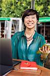 Woman with black hair wearing green shirt sitting in front of laptop at table in a street cafe, holding digital tablet, smiling at camera.