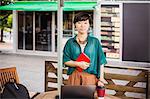 Woman with black hair wearing green shirt standing outdoors at a street cafe, looking at camera.