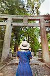 Rear view of young woman wearing blue dress and hat standing at Shinto Sakurai Shrine, Fukuoka, Japan.