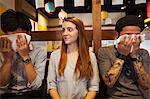 Three smiling people, woman and two men, sitting side by side at a table in a restaurant, men wiping their faces with wet towels.