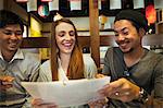 Three smiling people, woman and two men, sitting side by side at a table in a restaurant, looking at menu.