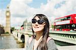 Smiling woman with black hair wearing sunglasses standing on Westminster Bridge over the River Thames, London, with the Houses of Parliament and Big Ben in the background.