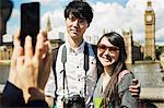 Smiling woman with black hair taking picture of couple with smartphone, standing on Westminster Bridge over the River Thames, London, with the Houses of Parliament and Big Ben in the background.