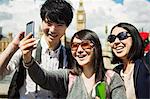 Smiling man and two women with black hair taking selfie with smartphone, standing on Westminster Bridge over the River Thames, London, with the Houses of Parliament and Big Ben in the background.