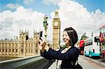 Smiling woman with black hair taking picture with smartphone, standing on Westminster Bridge over the River Thames, London, with the Houses of Parliament and Big Ben in the background.