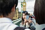 Smiling woman with black hair taking picture of couple with smartphone, standing on Westminster Bridge over the River Thames, London, with the Houses of Parliament and Big Ben in the background.