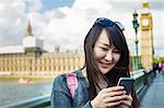 Smiling woman with black hair looking at smartphone, standing on Westminster Bridge over the River Thames, London, with the Houses of Parliament and Big Ben in the background.
