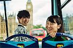 Smiling man and woman with black hair sitting at the front on the top of a Double-Decker bus in London, looking at camera, Houses of Parliament and Big Ben in the background.