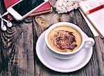 white cup with cappuccino behind the mobile phone with headphones on a gray wooden background