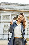 Stylish autumn in Paris. Portrait of happy young fashion-monger in trench coat near Arc de Triomphe in Paris, France showing victory gesture