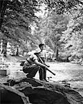 1940s 1950s FATHER AND BOY BY SIDE OF STREAM PICNIC BASKET MAN KNEELING HOLDING FISHING GEAR SUMMERTIME