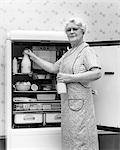 1930s 1940s SENIOR WOMAN HOUSEWIFE GRANDMOTHER LOOKING AT CAMERA TAKING TWO GLASS BOTTLES OF MILK OUT OF KITCHEN REFRIGERATOR