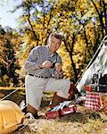 1960s MAN LOOKING AT CAMERA AT CAMPSITE ATTACHING LURE TO FISHING ROD