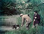 1950s MAN WOMAN COUPLE SHOTGUN GUNNING HUNTING BY LAKE POND BLACK LABRADOR DOG RETRIEVER RETURNING WATER FOWL DUCK