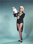 1960s BLOND WOMAN MAGICIAN WEARING BLACK CUTAWAY COAT COSTUME LOOKING AT CAMERA PULLING WHITE RABBIT OUT OF MAGIC TOP HAT