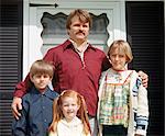 1970s SINGLE FATHER WITH HIS ARM AROUND HIS CHILDREN LOOKING AT CAMERA STANDING BY FRONT DOOR OF HOUSE