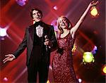 1970s WOMAN AND MAN WITH MICROPHONE SINGING AND GESTURING STROBE LIGHTS IN BACKGROUND TUXEDO GOWN ENTERTAINING