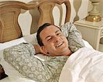1950s 1960s SMILING MAN LYING IN BED WEARING PAJAMAS LOOKING AT CAMERA