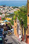 Overview of city viewed from traditional cooblestone street with ATV and woman walking on sidewalk in San Miguel de Allende, Mexico