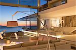 Illuminated, modern luxury home showcase interior living room with ocean view at dusk