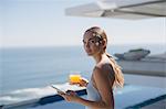 Portrait woman using digital tablet and drinking orange juice on sunny luxury patio with ocean view