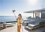 Woman in bathing suit taking selfie with selfie stick on sunny modern, luxury home showcase exterior patio with ocean view