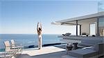 Woman practicing yoga mountain pose on sunny modern, luxury home showcase exterior patio with ocean view