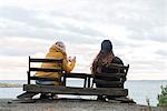 Rear view of teenage girl and young woman sitting on wooden chairs by lake and taking pictures