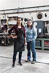 Two women wearing apron and Denim shirt standing in metal workshop, smiling at camera.