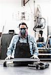 Woman wearing safety glasses and dust mask standing in metal workshop, looking at camera.