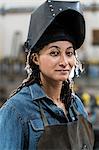 Portrait of woman wearing apron and welding mask standing in metal workshop, smiling at camera.