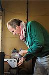 A craftsman in a knife makers workshop, holding a kitchen knife and shaping and finishing the wooden handle with a surface grinder a rotating wheel.