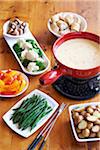 Cheese fondue in red pot with dishes of vegetables and bread on a wooden table