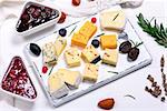 square pieces of camembert cheese, roquefort, cheddar and brie on a white wooden board, next to a raspberry and strawberry jam, top view