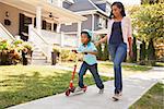 Mother Walks With Son As He Rides Scooter Along Sidewalk