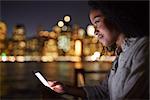 Woman Uses Mobile Phone At Night With City Skyline In Background