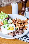 Beef rump steak topped with fried egg and chives herbs served with golden chips and fresh salad on a white plate on a wooden table