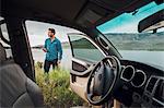 Mid adult man standing beside Dillon Reservoir, holding smartphone, view through parked car, Silverthorne, Colorado, USA