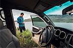 Mid adult man standing beside Dillon Reservoir, holding smartphone, view through parked car, Silverthorne, Colorado, USA