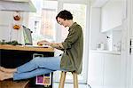 Mid adult woman sitting in kitchen, using laptop, laughing