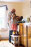 Girl on stool drinking while father prepares food in kitchen