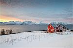 A traditional red house in the snowy landscape at sunset on the fjord, Djupvik, Lyngen Alps, Tromso, Norway, Europe