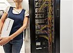 Female technician leaning against a server in a large computer server room, with a Network Security notebook under her arm.