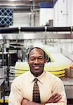 Portrait of an African American male management person standing next to a conveyor belt of lemon flavoured water in a bottling plant.
