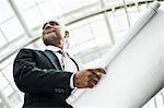 A black businessman holding plans for a new office space in a large glass covered walkway.