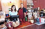 Caucasian woman and man, business owners of a coffee shop, at the counter. Displays of fresh baked goods.
