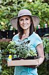 A woman in a sun hat carrying a tray of herbs and plants,  shopping at a garden centre nursery.